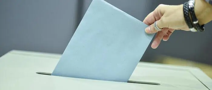 Das Bild zeigt eine Wahlurne, in die gerade ein Kuvert geworfen wird