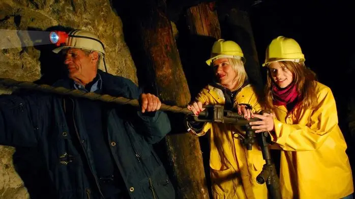 Im Schaustollen Knappenberg steht ein Mann mit Helm und Stirnleuchte, ihm folgen zwei Besucherinnen in gelben Jacken und gelben Schutzhelmen.