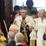 König Charles III. in königlichem Gewand mit Camilla an der Hand. Davor sitzen Personen.