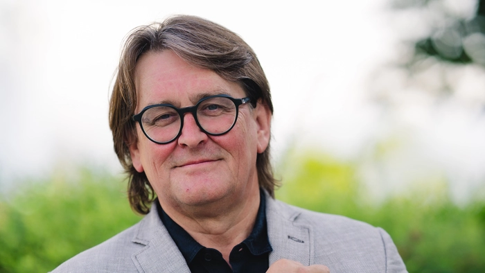 Ein Mann mit grauem Anzug, schwarzer Brille und leicht grauen Haaren blickt mit verkreuzten Händen verschmitzt in die Kamera