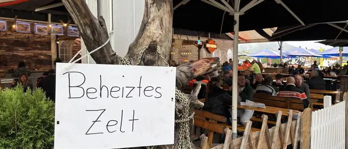 Auf einem Schild steht „beheiztes Zelt“, im Hintergrund sieht man Gäste auf Tischen sitzen