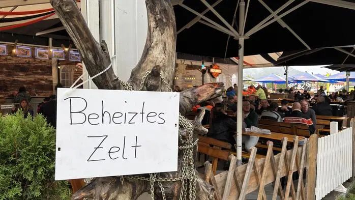 Auf einem Schild steht „beheiztes Zelt“, im Hintergrund sieht man Gäste auf Tischen sitzen