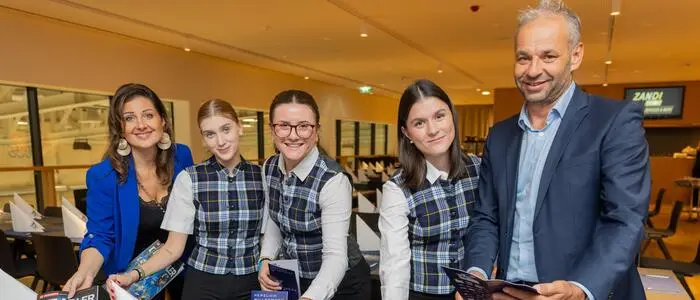 Eine Frau und ein Mann stehen mit drei Schülerinnen in gleicher Servicekleidung an einem gedeckten Tisch