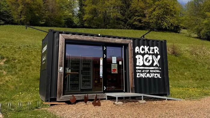 Ein Selbstbedienungsladen mit Glasfront steht auf einer Wiese, dahinter erheben sich Bäume