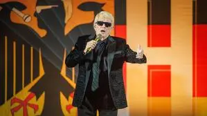 Heino, mit einem schwarzen Anzug und Sonnenbrille, hält ein Mikrofon in der Hand, er steht vor einer Deutschlandfahne