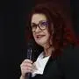 Claudia Schönbacher mit einem Mikrofon in der Hand. Sie hat lange rot gefärbte Locken, trägt eine Brille und dezente Kleidung in Schwarz und Weiß