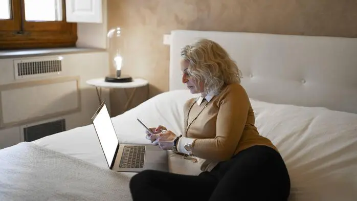 Eine Frau liegt in einem Hotelzimmerbett. Vor ihr steht ein offener Laptop, in der Hand hält sie ein Handy.