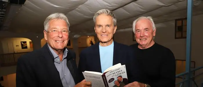 Drei gepflegte ältere Herren lachen in die Kamera und halten ein Buch