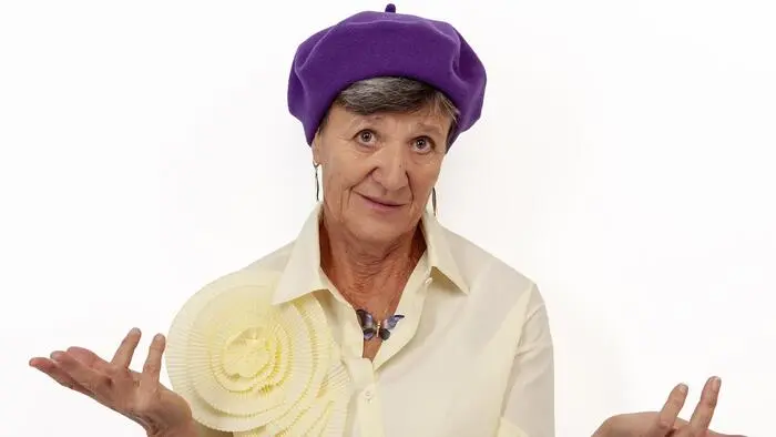 Eine ältere Dame schaut fragend in die Kamera und hat einen auffälligen lilafarbenen Hut auf.