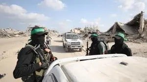 Vier Hamas-Terroristen sitzen bewaffnet und vermumt auf der Ladefläche eines Pickups. Dahinter fährt ein weiteres weißes Fahrzeug und die zerstörten Gebäude im Gazastreifen sind zu erkennen.