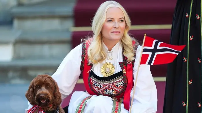 Kronprinzessin Mette-Marit sitzt in traditioneller Kleidung. Ihre rechte Hand liegt auf einem Hund, links hält sie die norwegische Flagge.