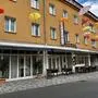 Hausfassade mit der Aufschrift „Delphi“, davor einige Tische mit Sesseln. Zur Dekoration hängen bunte Regenschirme in der Gasse