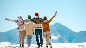 Four happy friends are standing and embracing against snow capped mountains at sunny day. Winter vacations concept
