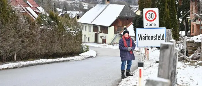Eine ältere Dame steht neben einer Ortstafel, auf der Weitensfeld steht. Es liegt Schnee um sie herum