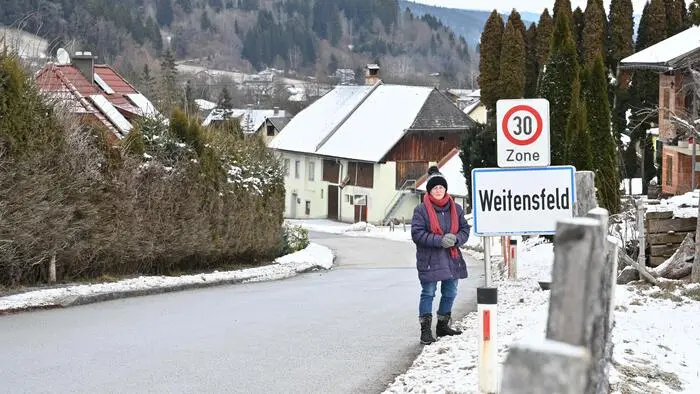 Eine ältere Dame steht neben einer Ortstafel, auf der Weitensfeld steht. Es liegt Schnee um sie herum