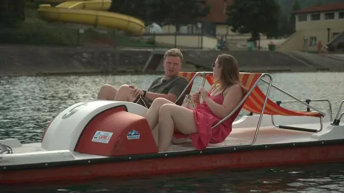 Eine Frau und ein Mann sitzen in sommerlicher Kleidung in einem Tretboot. Dahinter befindet sich eine Wasserrutsche