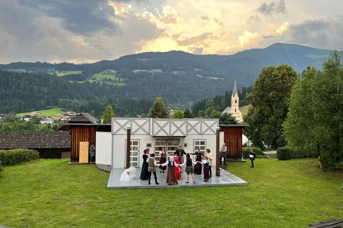 Gespielt wird in der Holzarena Arriach, vor der malerischen Kulisse der Nockberge, im Hintergrund die evangelische Kirche des Ortes und der Mittagskogel.