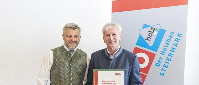Zwei Männer lächeln in die Kamera. Der Mann rechts hat eine Urkunde in der Hand