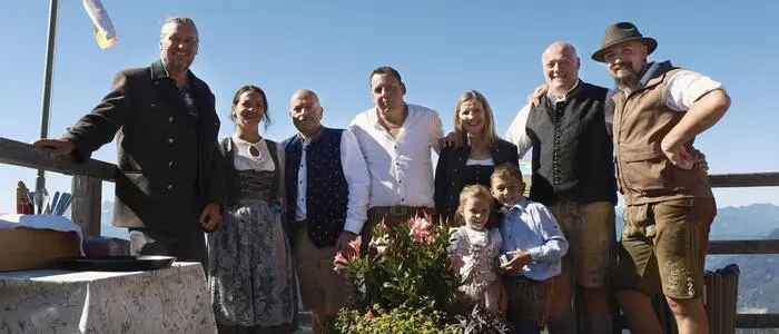 Eine Gruppe von Männern und Frauen in Tracht steht auf der Terrasse einer Almhütte