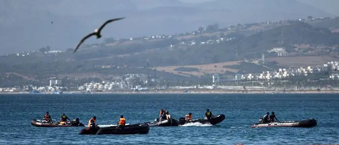 Mehrere Schlauchboote mit Einsatzkräften fahren auf dem Meer vor einer bergigen Küstenlandschaft. Im Vordergrund ist eine schwimmende orangefarbene Sperre zu sehen, über den Booten fliegt ein Vogel.
