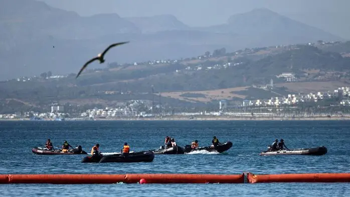 Mehrere Schlauchboote mit Einsatzkräften fahren auf dem Meer vor einer bergigen Küstenlandschaft. Im Vordergrund ist eine schwimmende orangefarbene Sperre zu sehen, über den Booten fliegt ein Vogel.