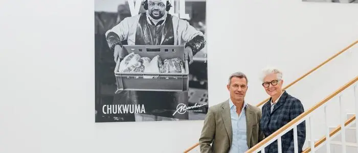 Ein Mann und eine Frau stehen auf einer Treppe mit einem großen Bild  eines Mannes mit einer Kiste voll Fleisch im Hintergrund