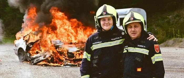 Ein junger und ein älterer Feuerwehrmann stehen vor einem brennenden Fahrzeug.