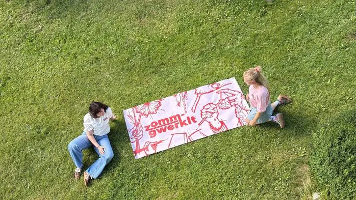 Ein großes Plakat liegt auf einer Wiese, zwei Frauen sitzen links und rechts davon