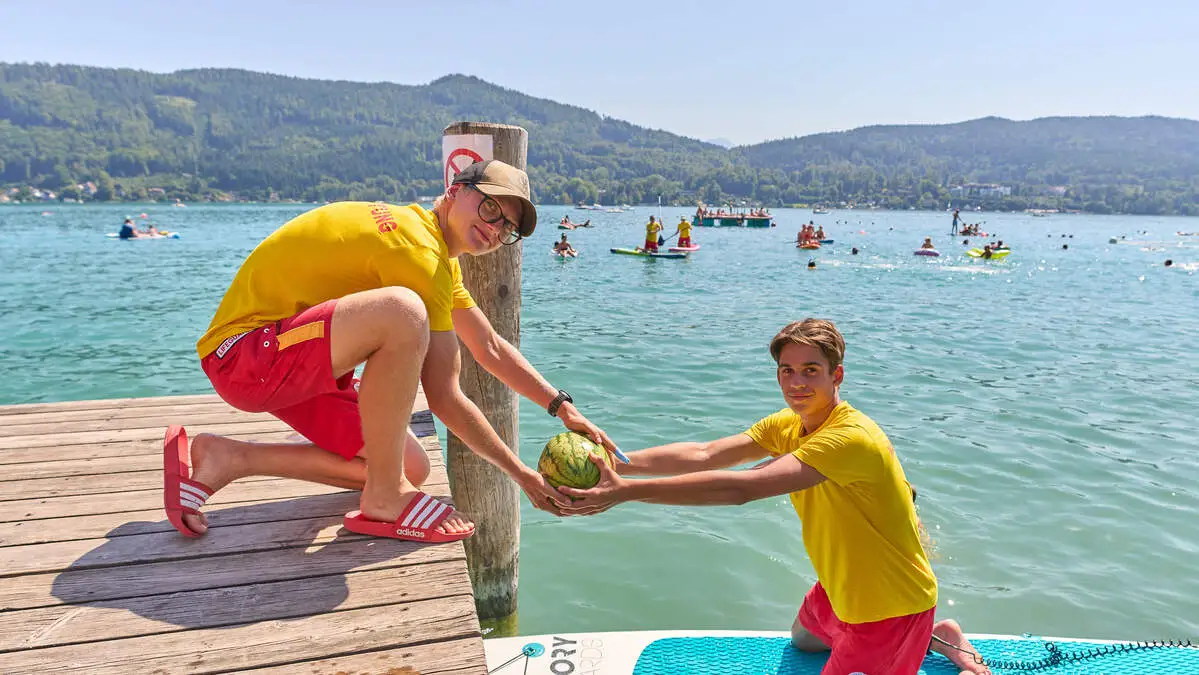 Ein Wasserretter hebt eine Melone von seinem SUP in die Hände eines Kollegen am Steg | Die Wassermelonen-Ernte in Krumpendorf war wieder ein voller Erfolg