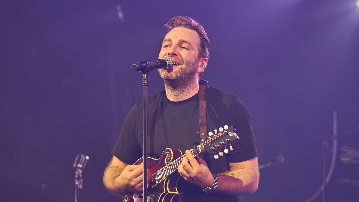 Ein Mann singt ins Mikrofon und spielt dabei Gitarre. | Josh kommt 2026 ins Schloss Albeck