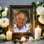 Ein älterer Mann lacht aus einem Bilderrahmen in die Kamera, drumherum sind weiße Rosen und Kerzen