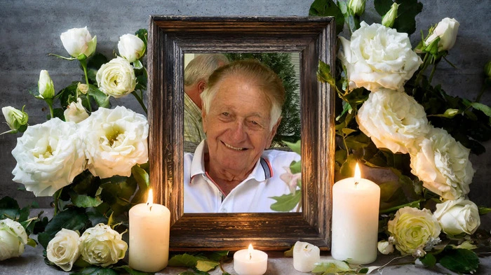 Ein älterer Mann lacht aus einem Bilderrahmen in die Kamera, drumherum sind weiße Rosen und Kerzen