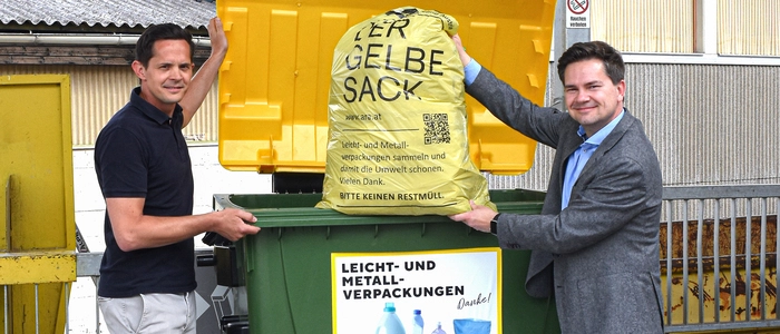 Zwei Männer werfen einen Gelben Sack in einen großen Container