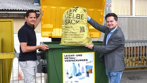 Zwei Männer werfen einen Gelben Sack in einen großen Container