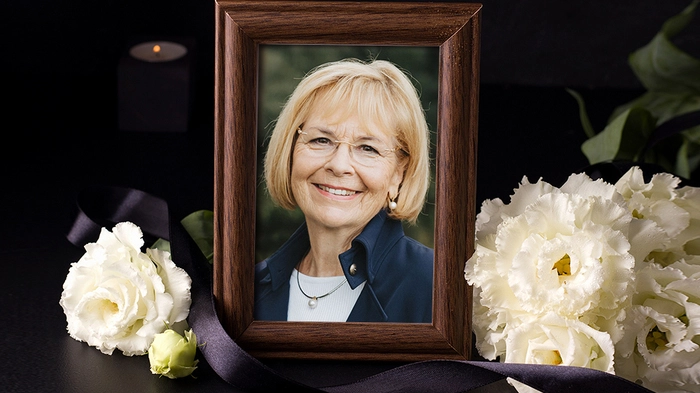 Eine blonde Frau mit Brille lacht in die Kamera, ihr Foto ist in einem Bilderrahmen umgeben von Blumen