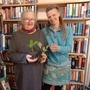 Zwei Frauen mit Pflanzen in der Hand, im Hintergrund sieht man Regale voller Bücher