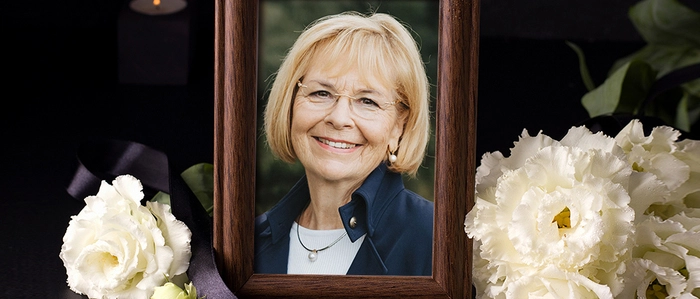 Eine blonde Frau mit Brille lacht in die Kamera, ihr Foto ist in einem Bilderrahmen umgeben von Blumen
