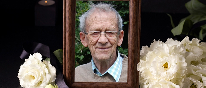 Ein Mann mit Brille in einem Bilderrahmen lacht in die Kamera, um den Bilderrahmen herum sind weiße Blumen