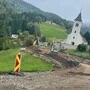 Eine aufgerissene Straße auf der Baustellenfahrzeuge stehen, im Hintergrund sieht man eine Grünfläche und Kirche