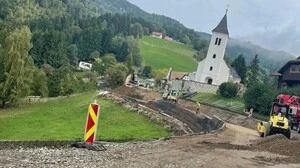 Eine aufgerissene Straße auf der Baustellenfahrzeuge stehen, im Hintergrund sieht man eine Grünfläche und Kirche
