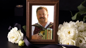 Ein Mann mit Harmonika in der Hand lacht in die Kamera, sein Bild ist in einem Bilderrahmen, umgeben von weißen Blumen