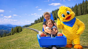 Eine Frau und ein Kind sitzen in einer Rodel, daneben steht ein gelbes Maskottchen in Menschengröße