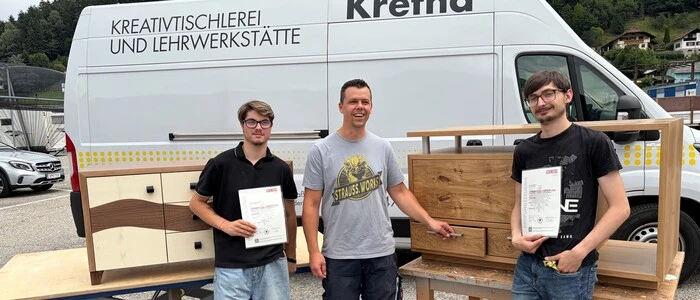 Drei Männer stehen vor Werkstücken aus Holz und einem Fahrzeug