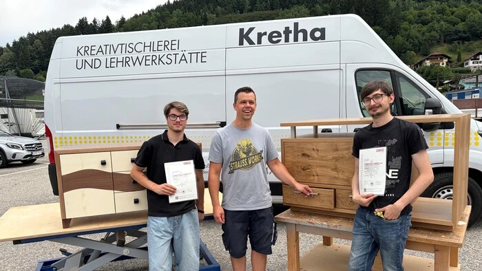Drei Männer stehen vor Werkstücken aus Holz und einem Fahrzeug