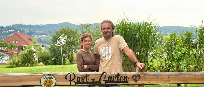 Zwei Menschen stehen hinter einem Gartenzaun mit der Aufschrift „RastGarten“