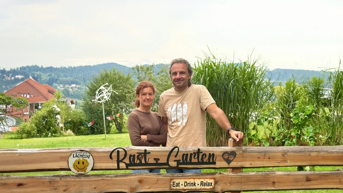 Zwei Menschen stehen hinter einem Gartenzaun mit der Aufschrift „RastGarten“