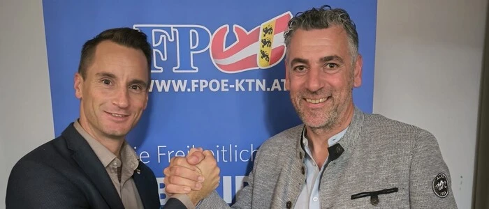 Zwei Männer geben sich die Hand und lächeln in die Kamera. Im Hintergrund sieht man ein FPÖ-Banner.