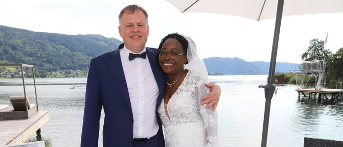 Ein Ehepaar lächelt bei seiner Hochzeit in die Kamera. Im Hintergrund sieht man den Ossiacher See.
