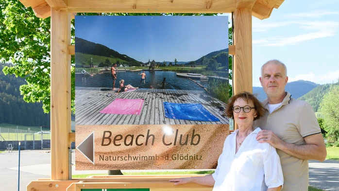 Zwei Menschen stehen neben einer Holztafel mit der Aufschrift „Beach Club“