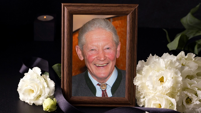 Ein gerahmtes Foto eines Mannes mit einer schwarzen Schleife, daneben weiße Rosen.